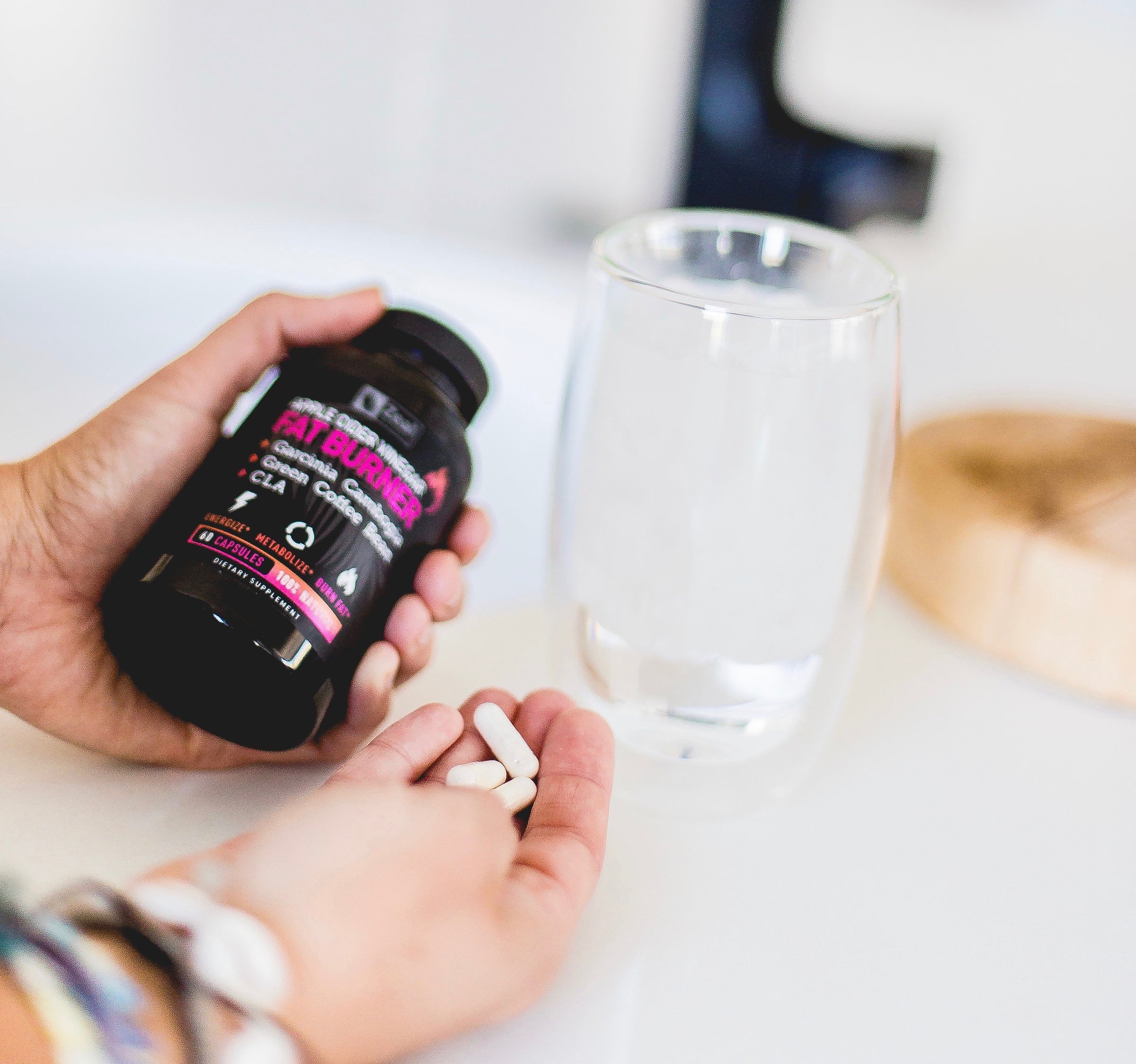 hands holding bottle of Fat burner in left hand, and pills in the right hand next to a glass of water